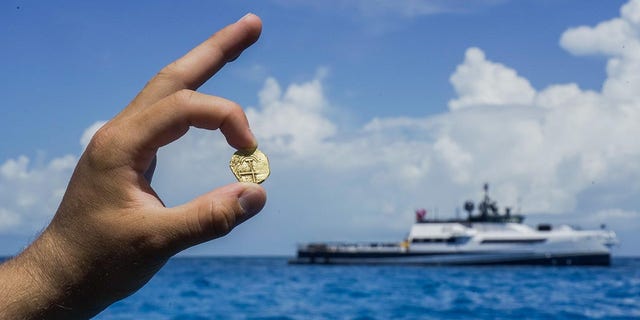 Um explorador segura uma moeda de ouro encontrada nas Bahamas, onde o barco de exploração de Allen pode ser visto de longe.