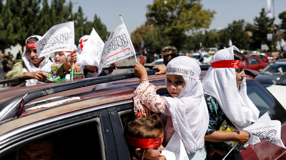 Meninas afegãs carregam as bandeiras do Emirado Islâmico enquanto participam do primeiro aniversário da retirada das forças americanas do Afeganistão
